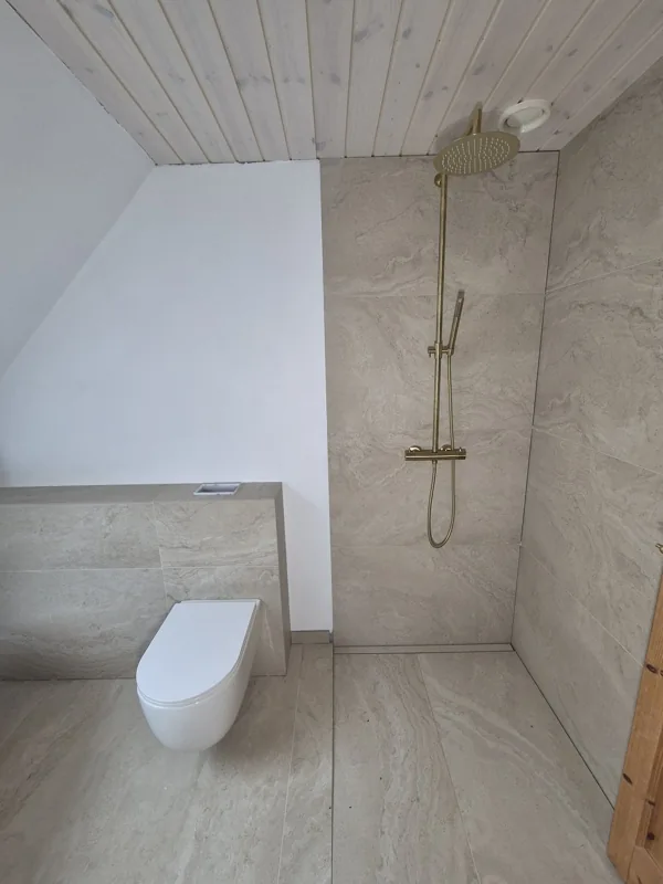 Væghængt toilet og bruseplads med messingfarvet armatur og store lyse fliser.